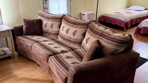 Ein Zimmer mit einem braunen Sofa, einem Bett mit gemustertem Überwurf, einer Lampe und einem Fenster mit Jalousien.