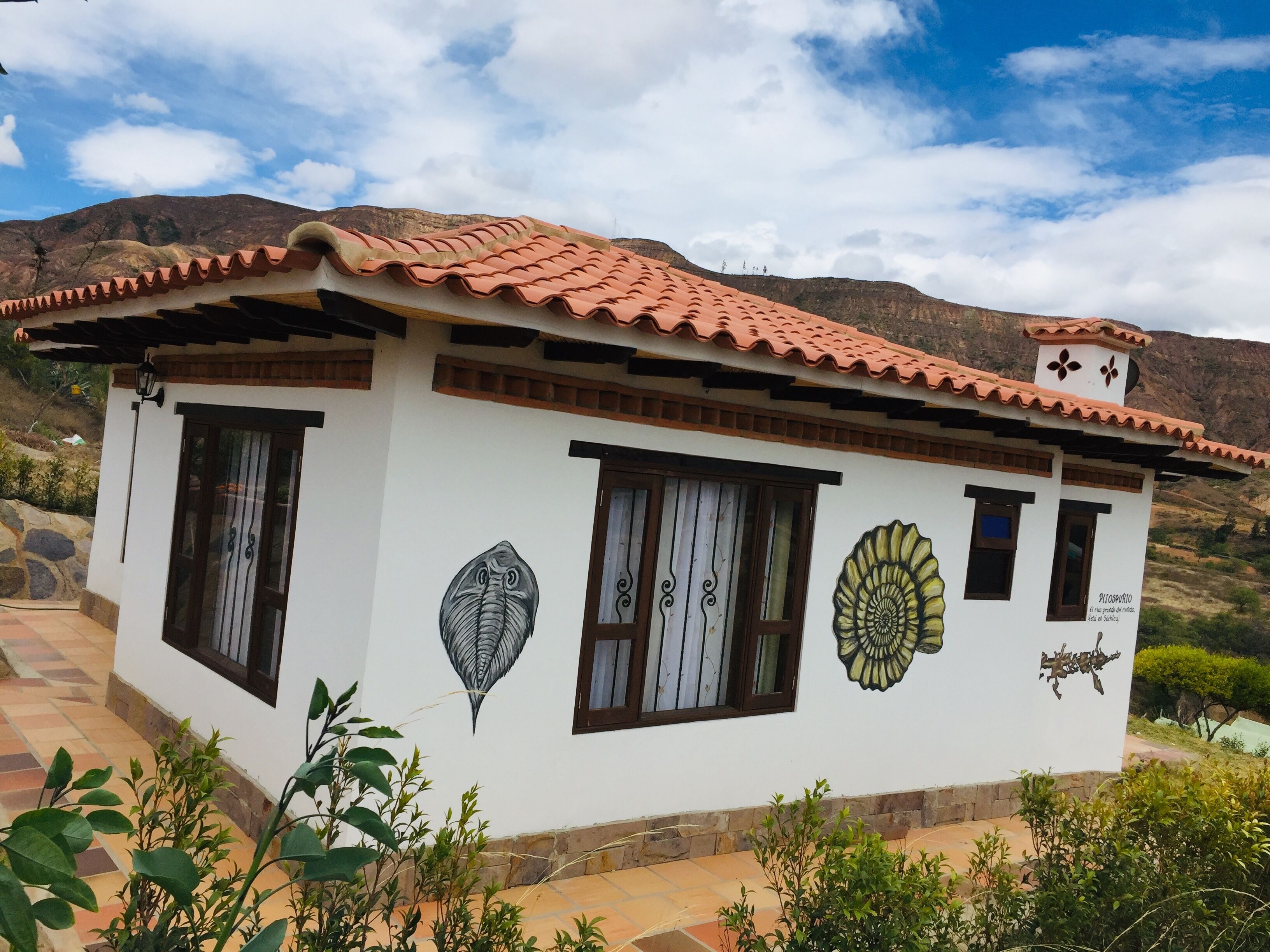 A white building with a red-tiled roof, featuring decorative wall art and a stone pathway leading to the entrance.