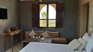 Ein Zimmer mit einem Bett, einem Holztisch mit Lampe, einer Couch mit Kissen, einem an der Wand befestigten Fernseher und einem Fenster mit Blick auf Grünflächen.
