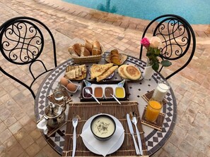 Une table ronde recouverte d’une nappe à carreaux, une théière, un bol de soupe, du pain et un verre de jus d’orange.