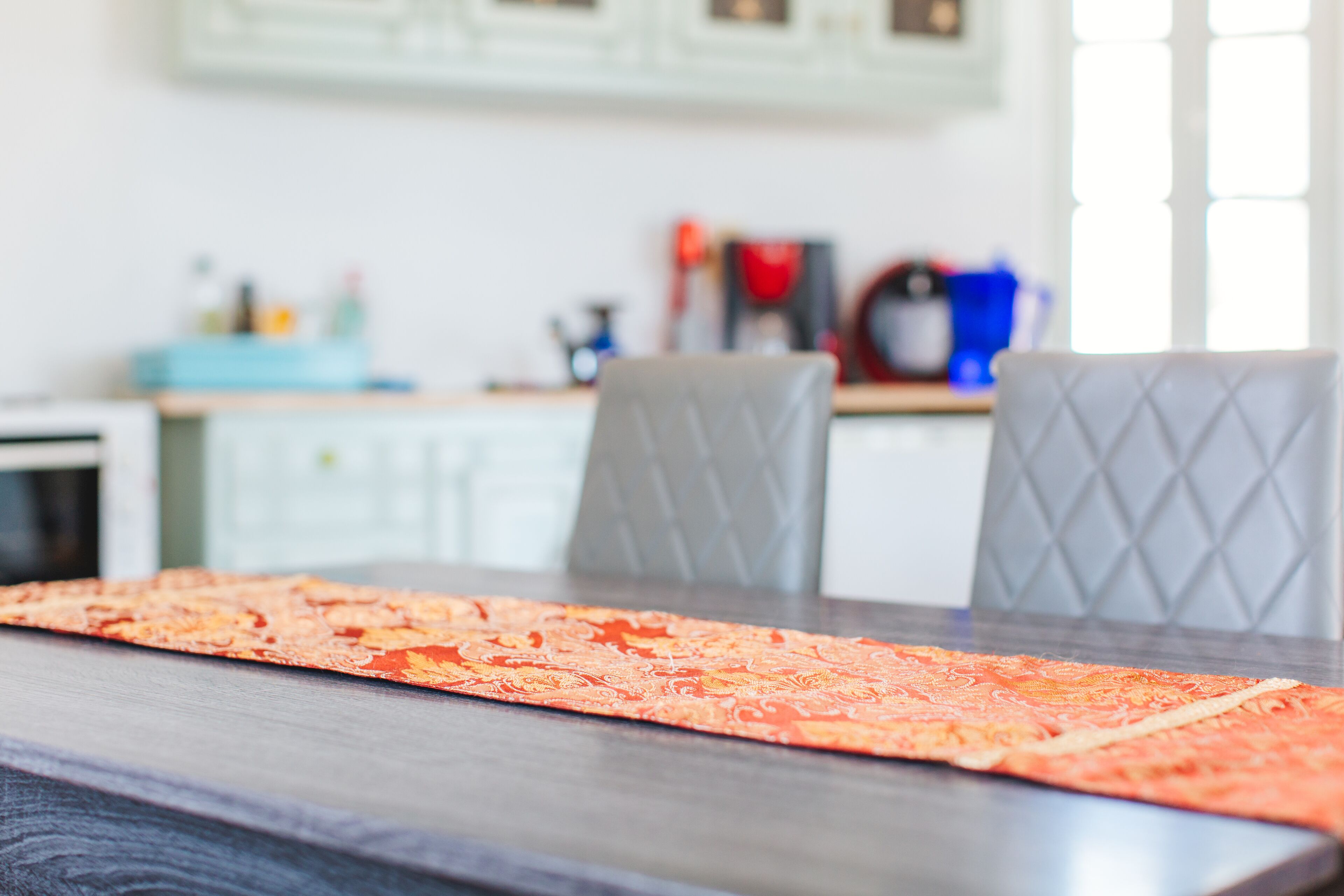 A dining table with a patterned table runner, a dark wooden table surface, and two grey upholstered chairs.