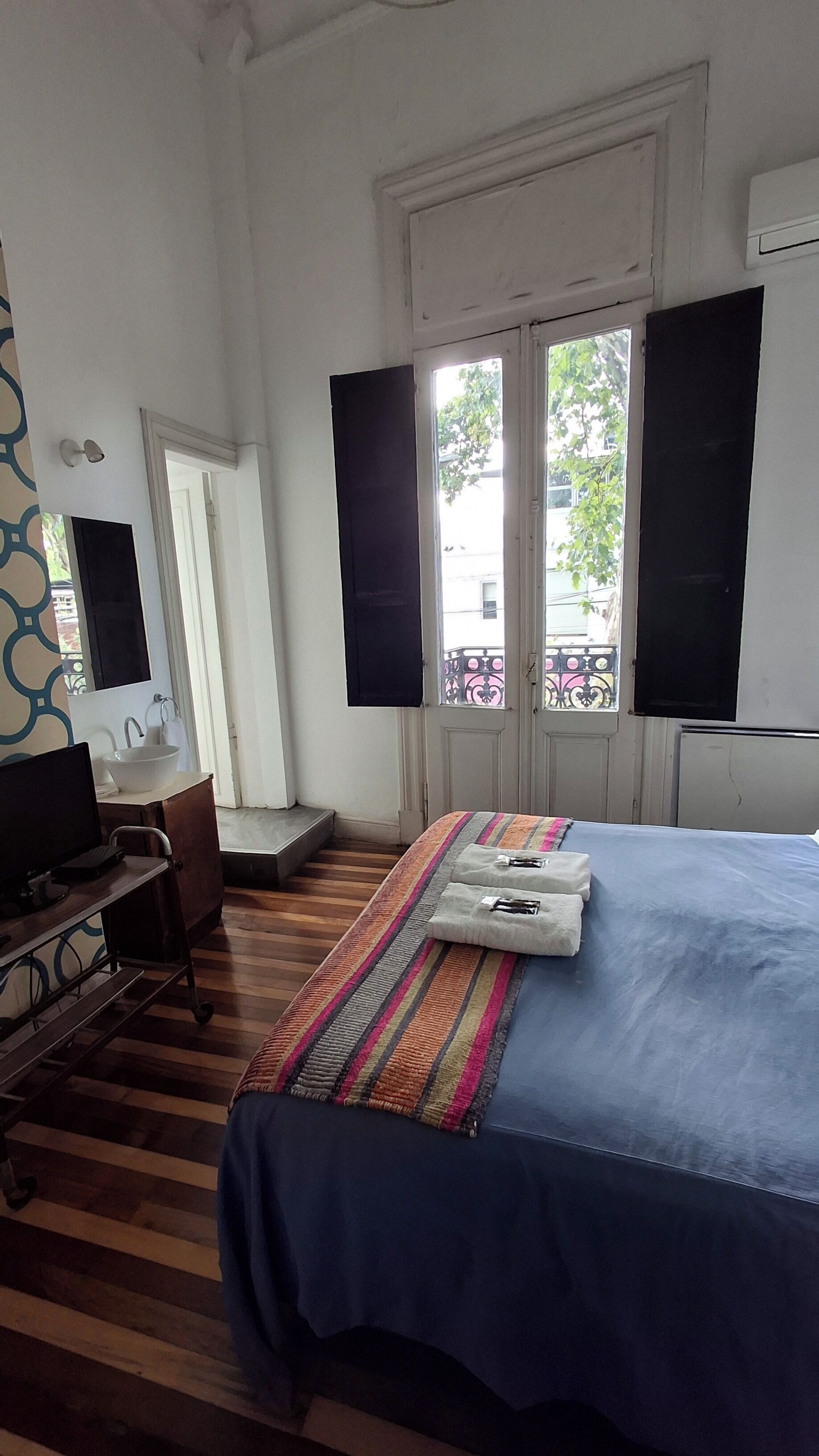 A bedroom with a bed, a desk with a computer, a window with shutters, and a view of the outside.