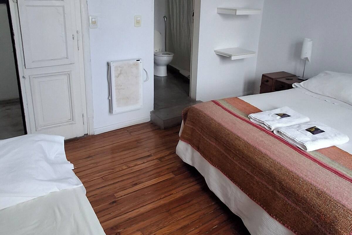 A hotel room with a wooden floor, a bed with a striped blanket, a white wardrobe, a shower area, and a small bedside table with a lamp.