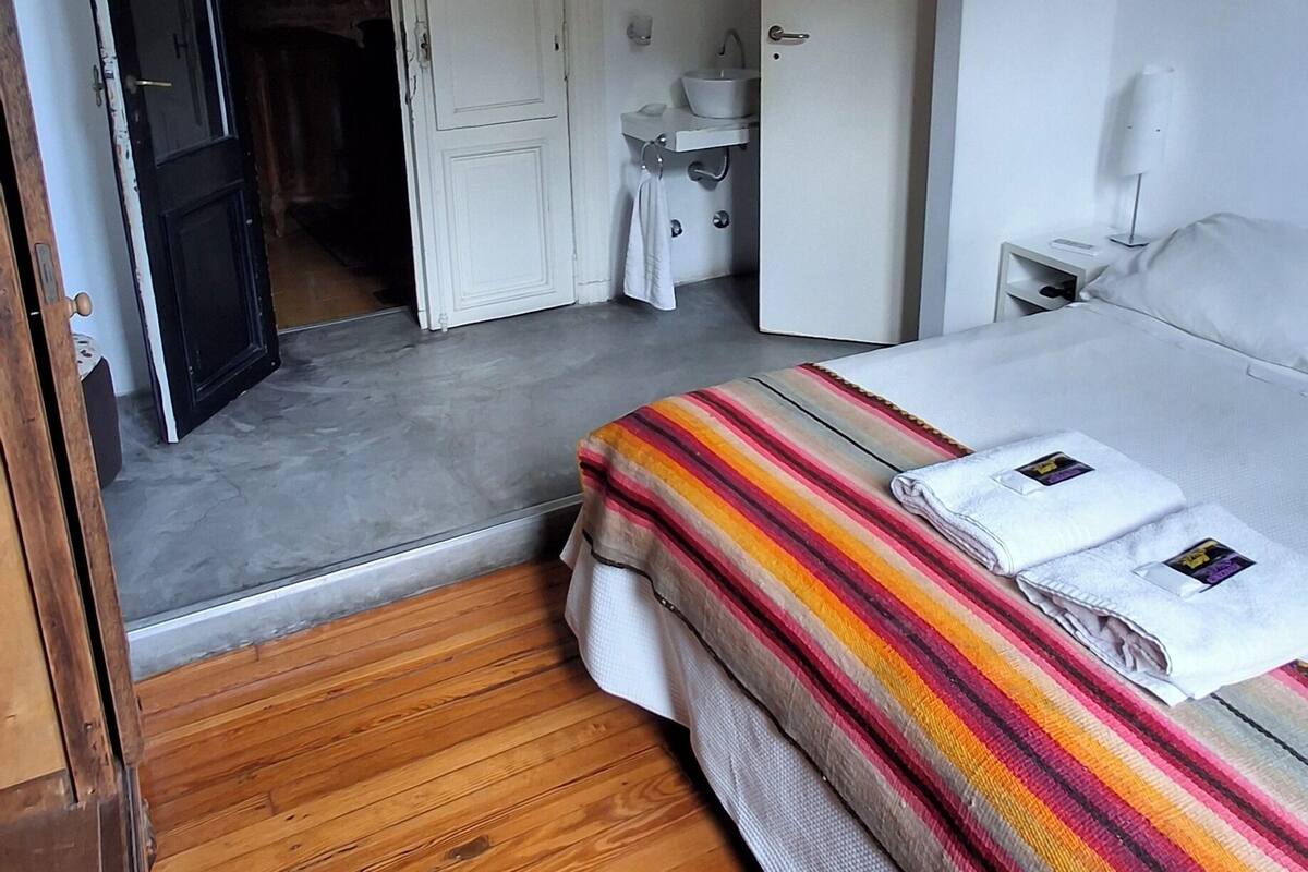 A bedroom with a bed, a wooden floor, a white wardrobe, a bathroom with a sink, and a door leading to another room.