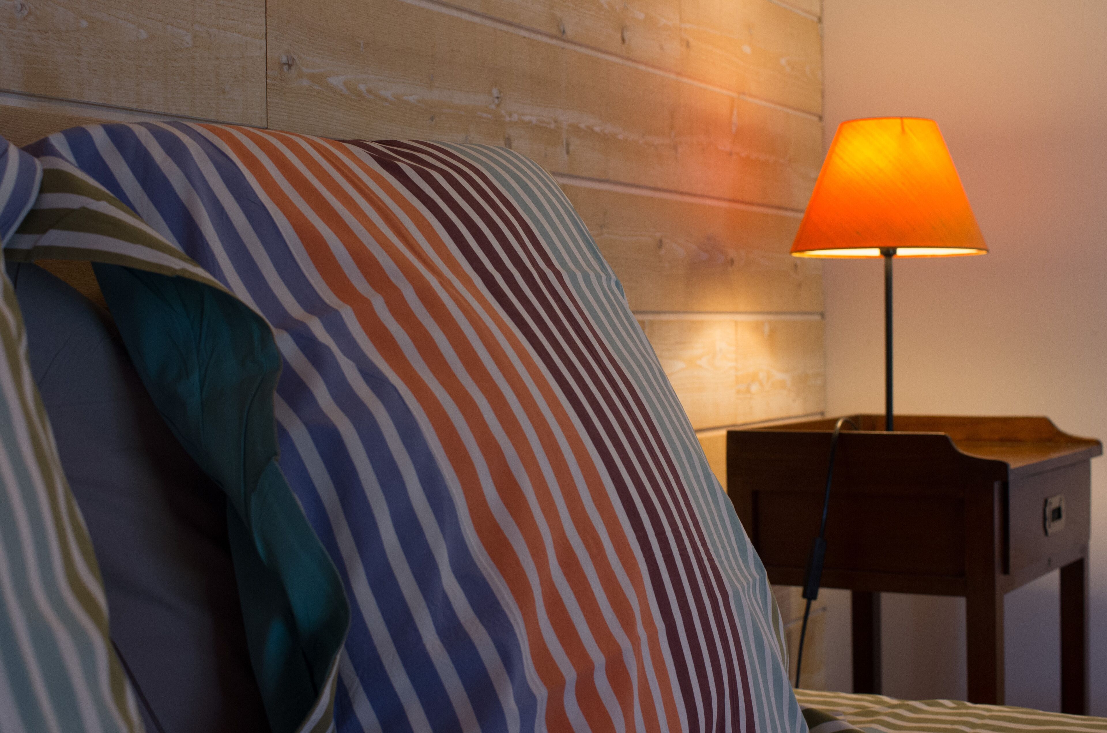 Una cama con almohadas a rayas y un cabecero de madera, una mesita de noche con una lámpara, y una pared de madera.