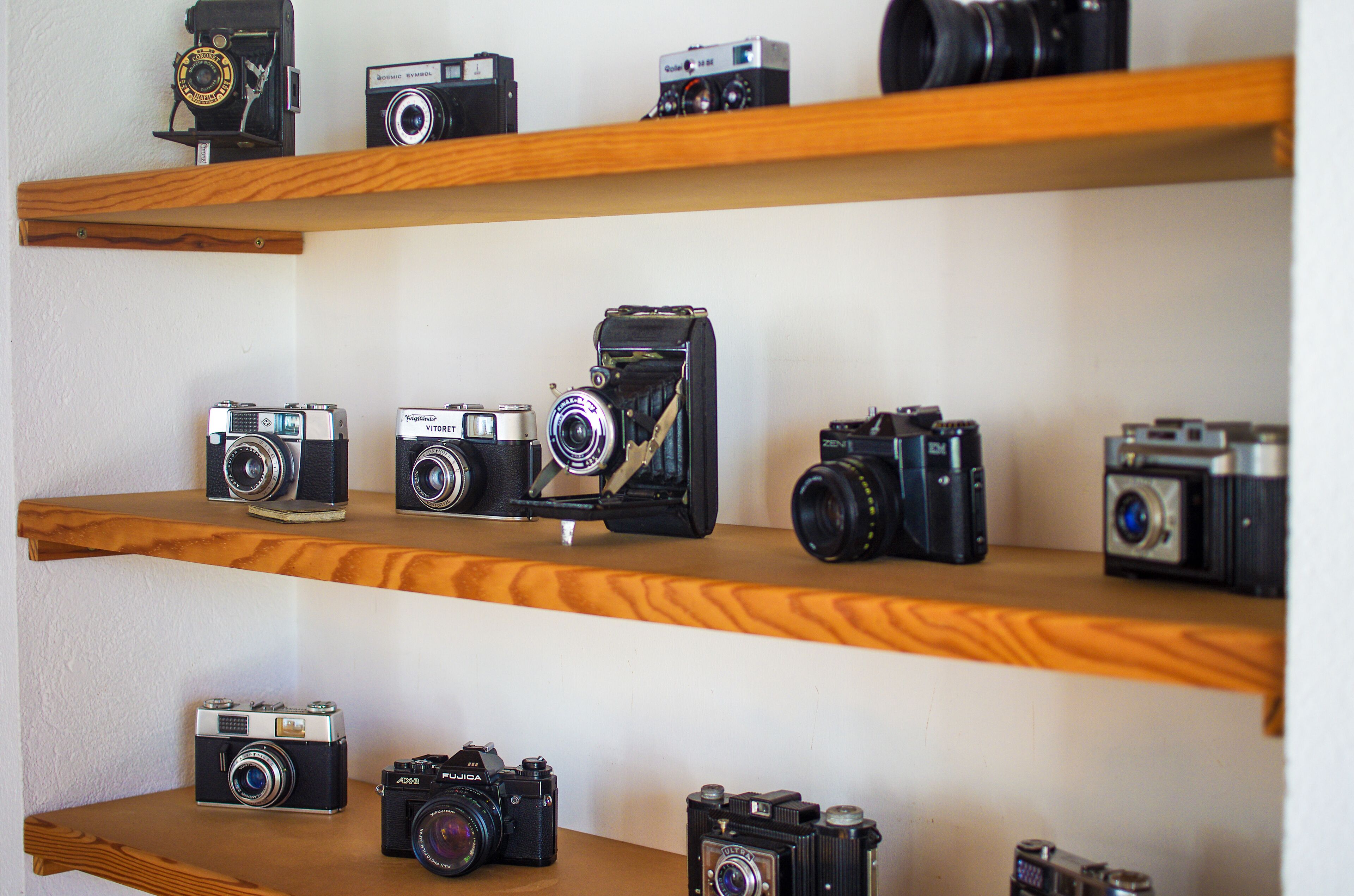A collection of vintage cameras displayed on wooden shelves against a plain wall.