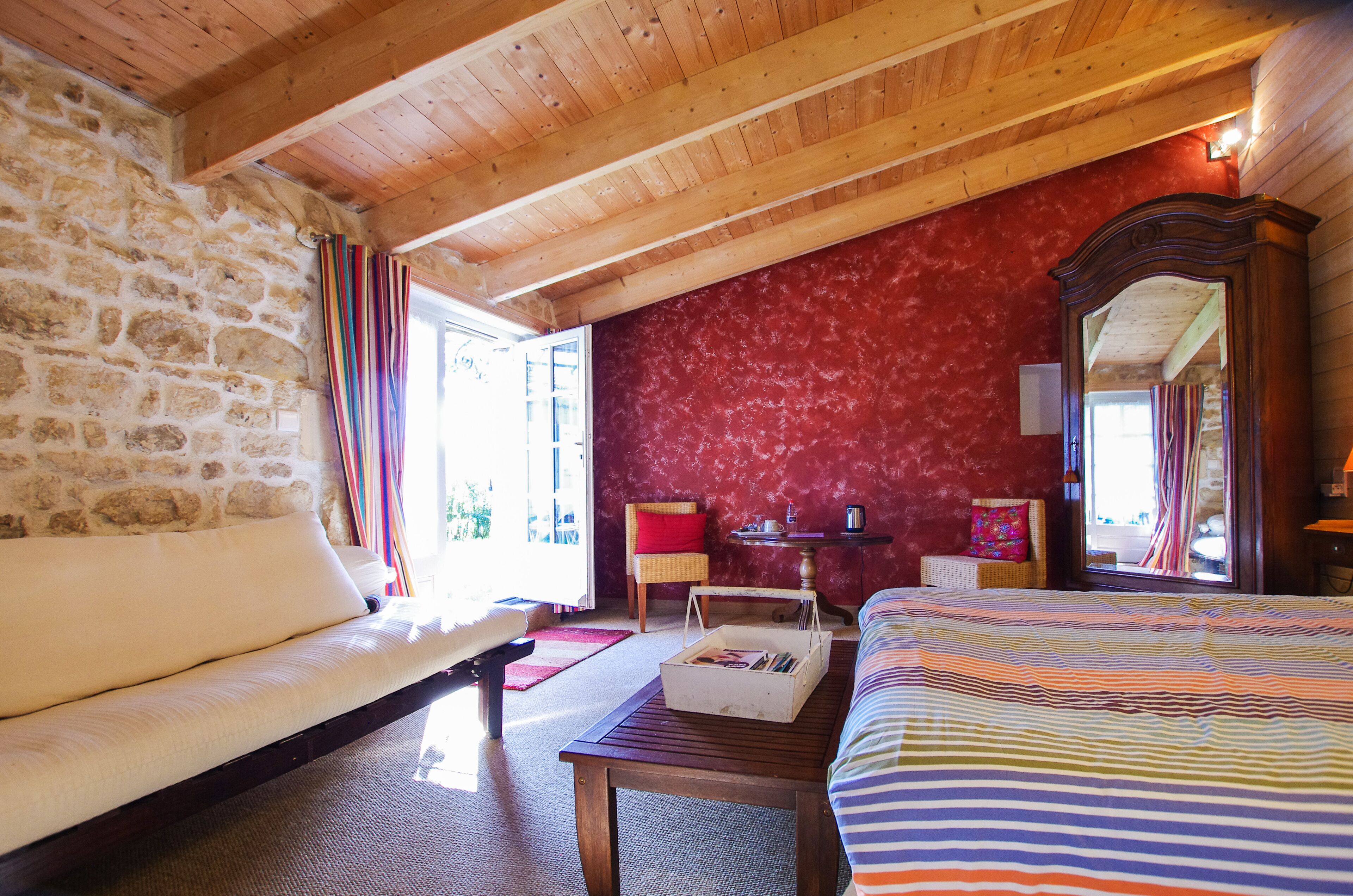 A cozy room with a stone wall, wooden ceiling beams, a red textured wall, a bed with striped bedding, a wooden table, a wicker chair, and a mirror.