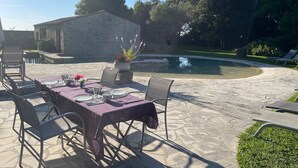 An outdoor patio with a stone table set for dining, surrounded by chairs, and a pool in the background.