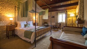 A bedroom with a four-poster bed, a wooden chair, a nightstand, and a stone wall.
