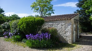 A stone building with a tiled roof, surrounded by lush greenery and purple flowers.
