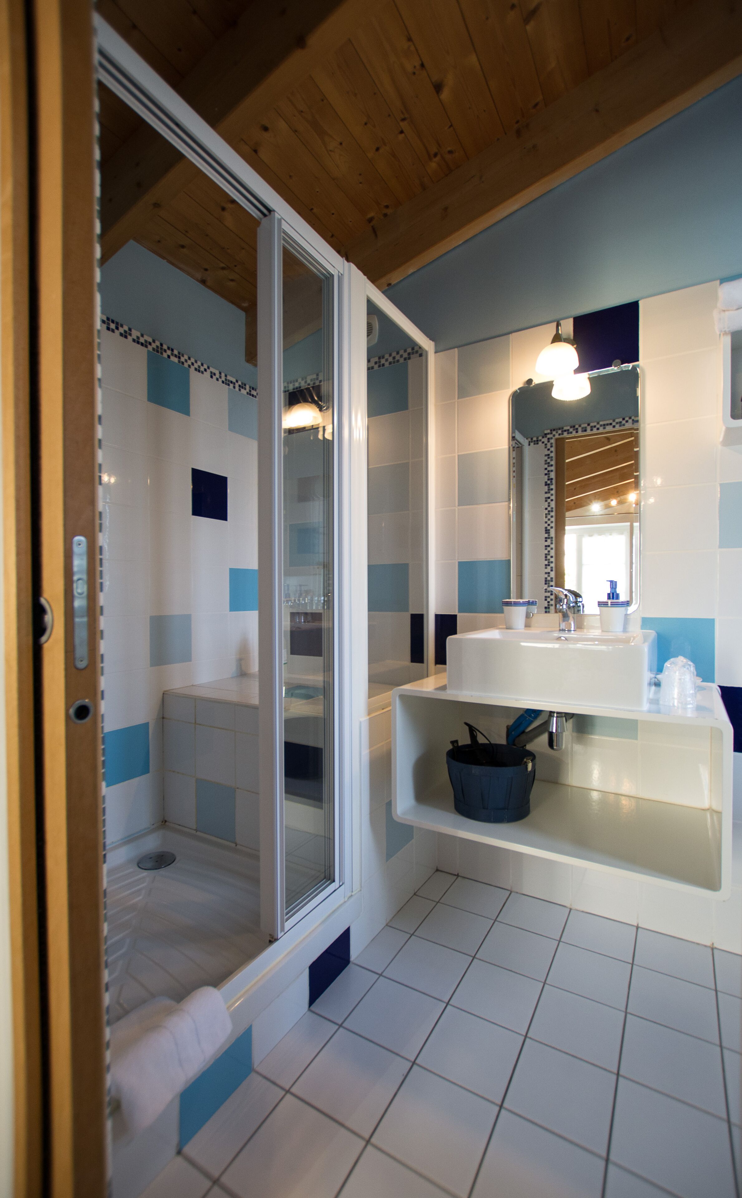 Un baño moderno con cabina de ducha de vidrio, un lavamanos blanco con dispensador de jabón azul y techo de madera.
