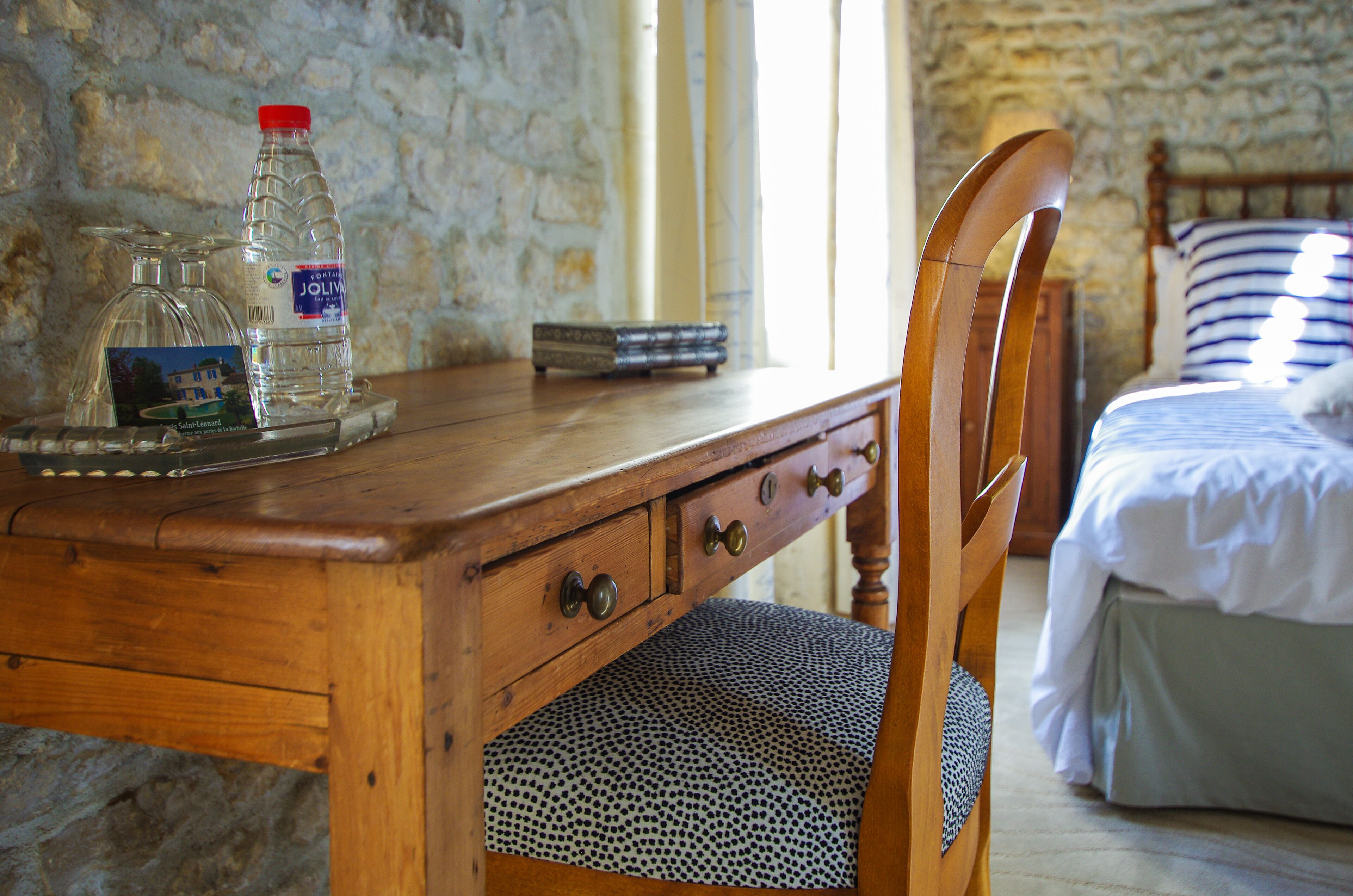 Un escritorio de madera con una botella de agua y un libro, una silla, y una cama con un almohadón a rayas en una habitación con paredes de piedra.