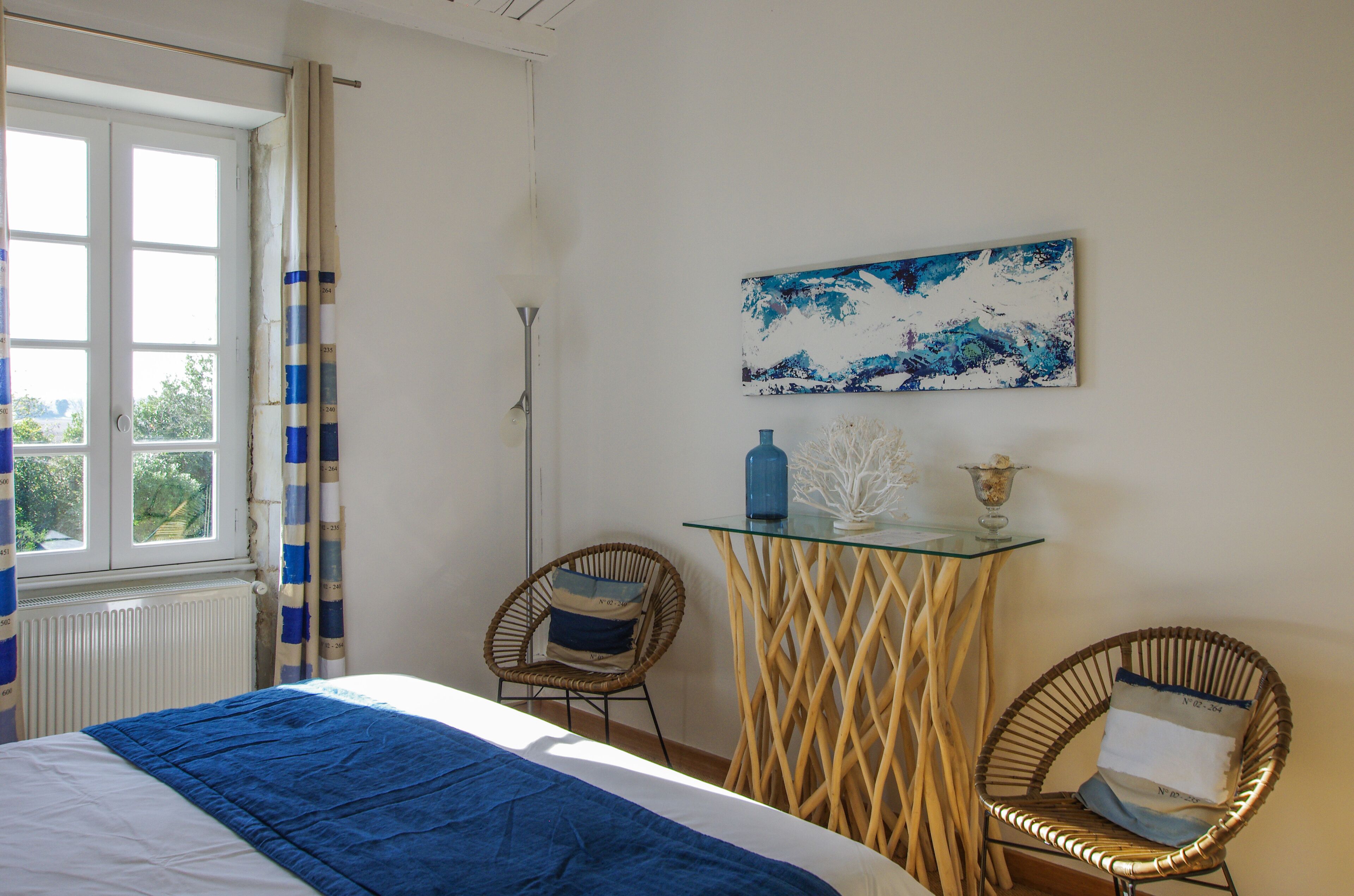 Un dormitorio con una cama, dos sillones de mimbre, una mesa de vidrio y un cuadro en la pared.