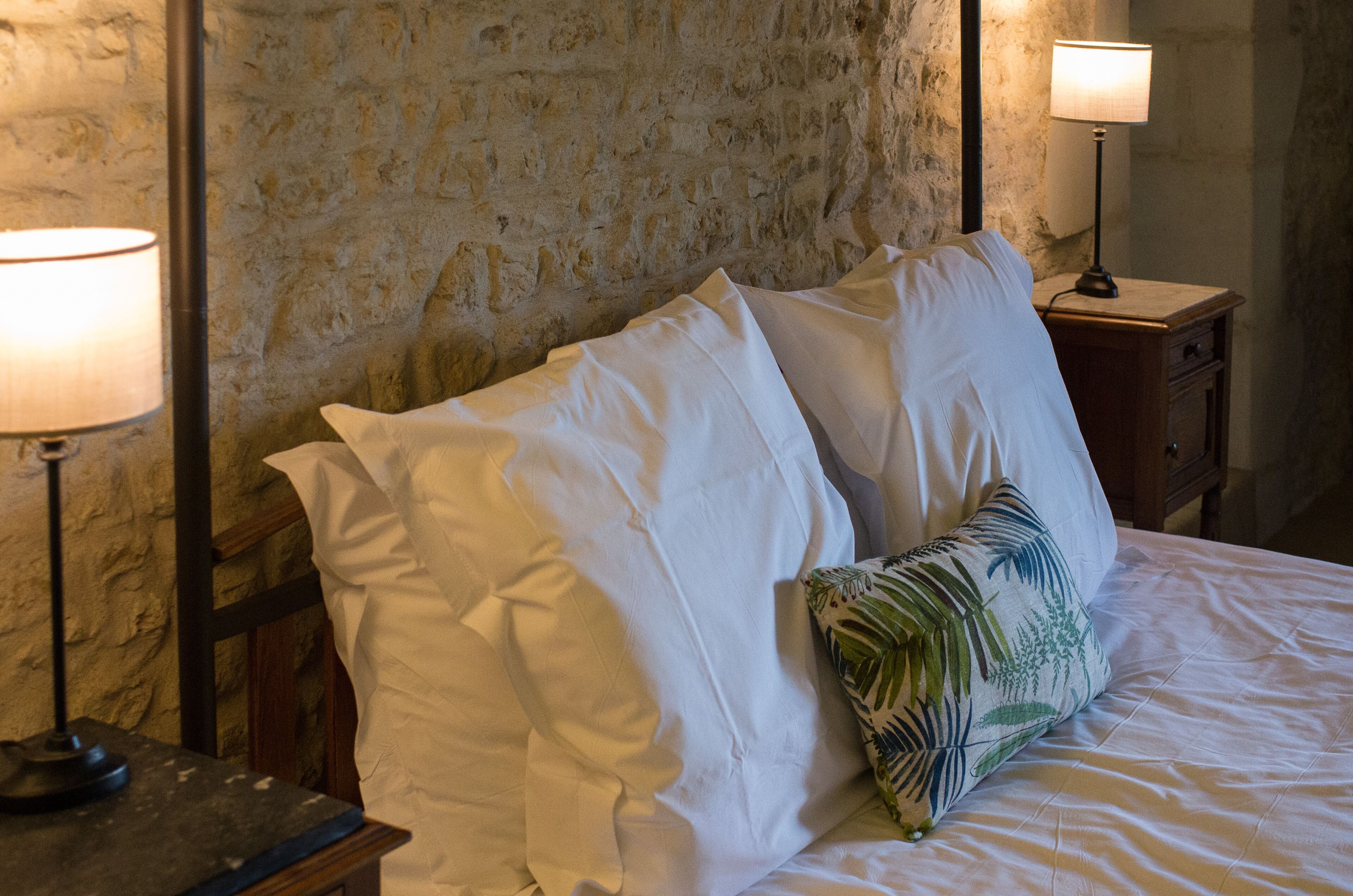 A bed with white linens and a palm leaf pillow, flanked by two bedside lamps on wooden nightstands, against a textured stone wall.