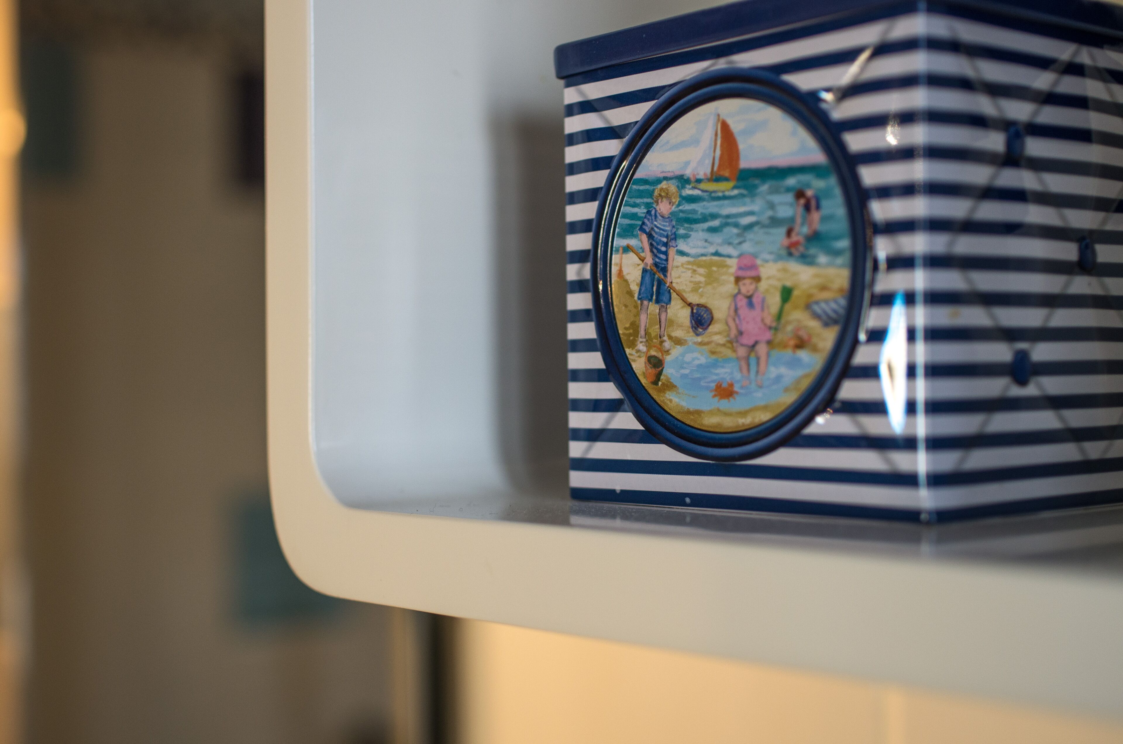 A striped container with a circular lid, featuring a beach scene with children and a sailboat.