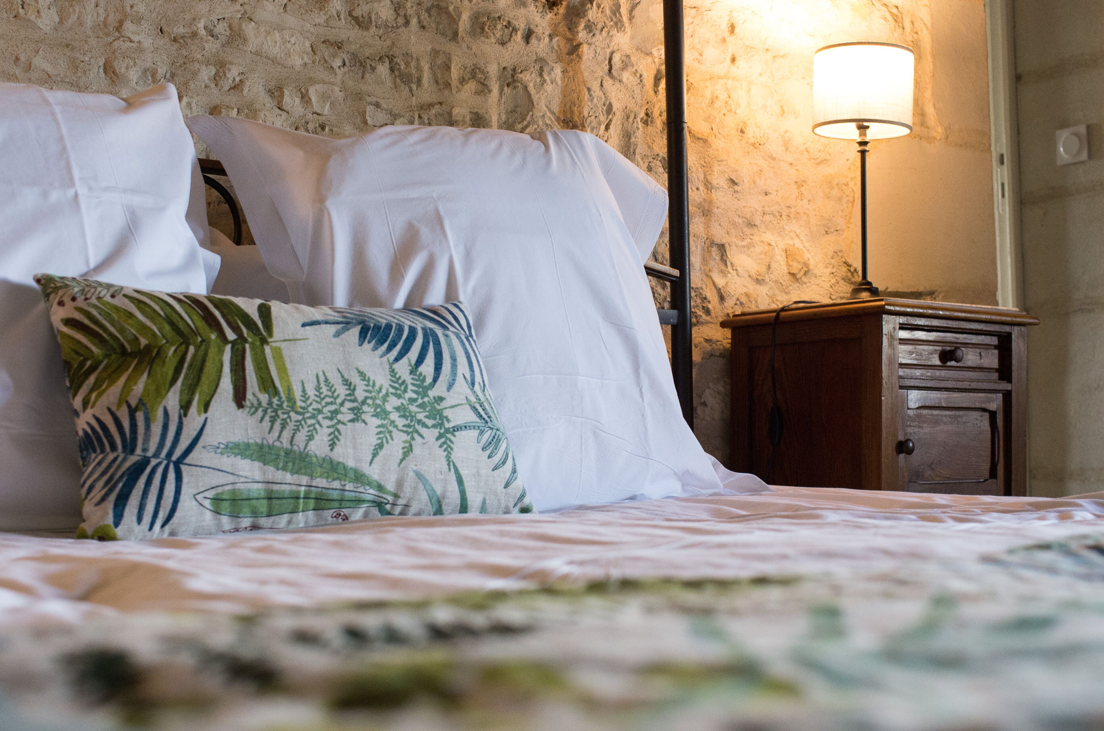 A bed with white linens, a green leaf-patterned pillow, and a wooden nightstand with a lamp.