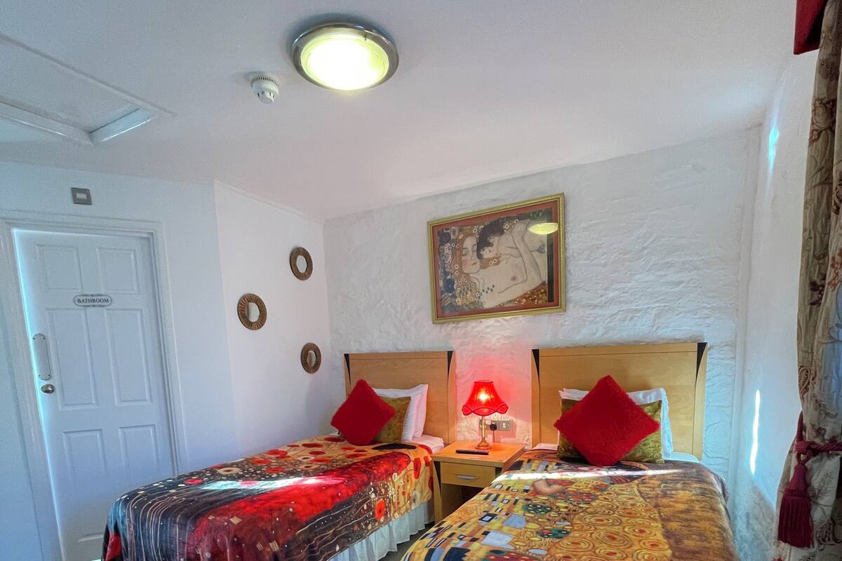 A hotel room with two beds, a painting on the wall, a ceiling light, and a door labeled "Bathroom".
