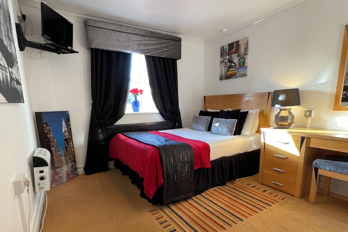 A hotel room with a bed, a desk, a chair, a radiator, a window with curtains, and a wall-mounted TV.