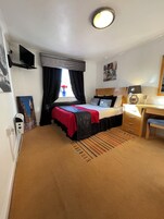 A hotel room with a bed, a desk, a chair, a radiator, a window with curtains, and a wall-mounted TV.