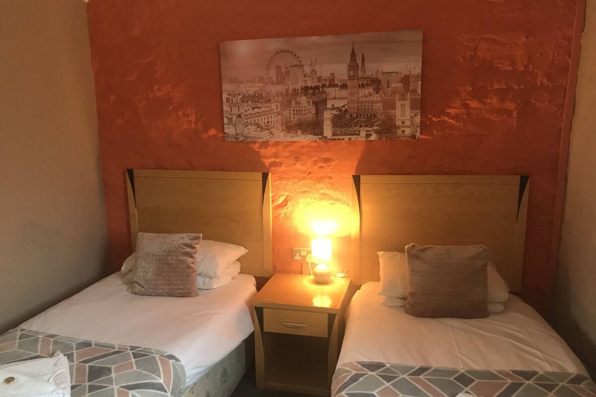 A hotel room with two beds, a nightstand with a lamp, and a cityscape painting on the wall.