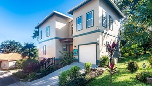 A two-story residential house with a garage, surrounded by lush greenery and a clear blue sky. - Tortuga Terrace-43 Lawson Rock 3 Bedroom Home by RedAwning (Roatan)