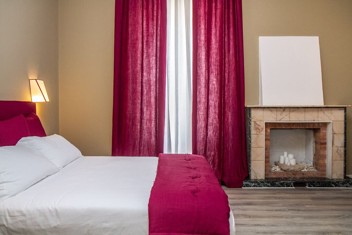 A hotel room with a bed, a red throw blanket, a fireplace, and red curtains.