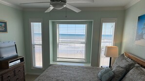 A bedroom with a bed, a television on a stand, a ceiling fan, and a view of the beach through the windows.