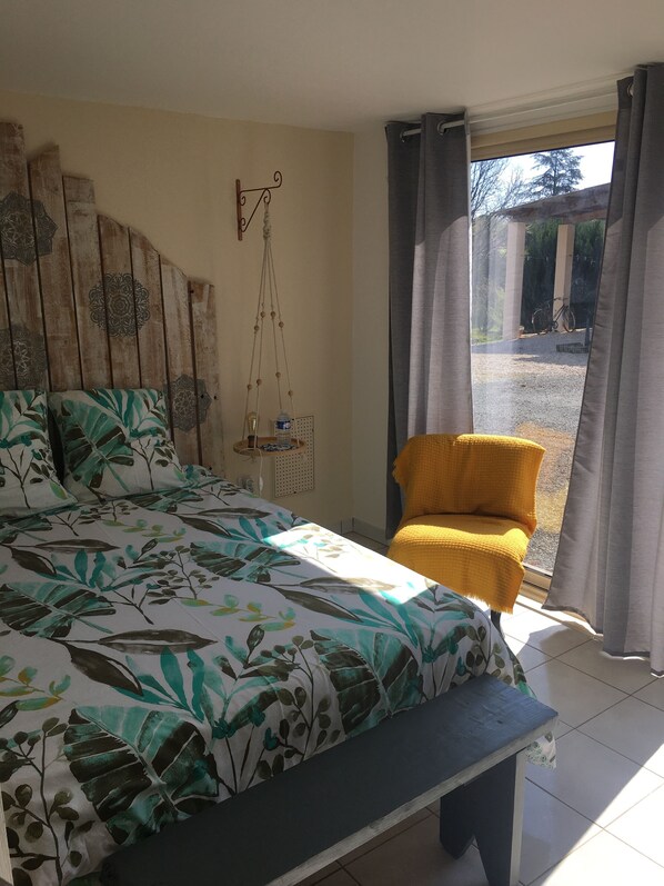 A bedroom with a bed, a bench, a chair, a hanging plant, and a view of the outside.