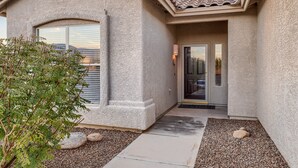 A stucco building with a tiled roof, a glass door entrance, and a small garden area with rocks and a tree. - Mystic Mirage is truly a desert oasis with pool and mountain views. (Tucson)