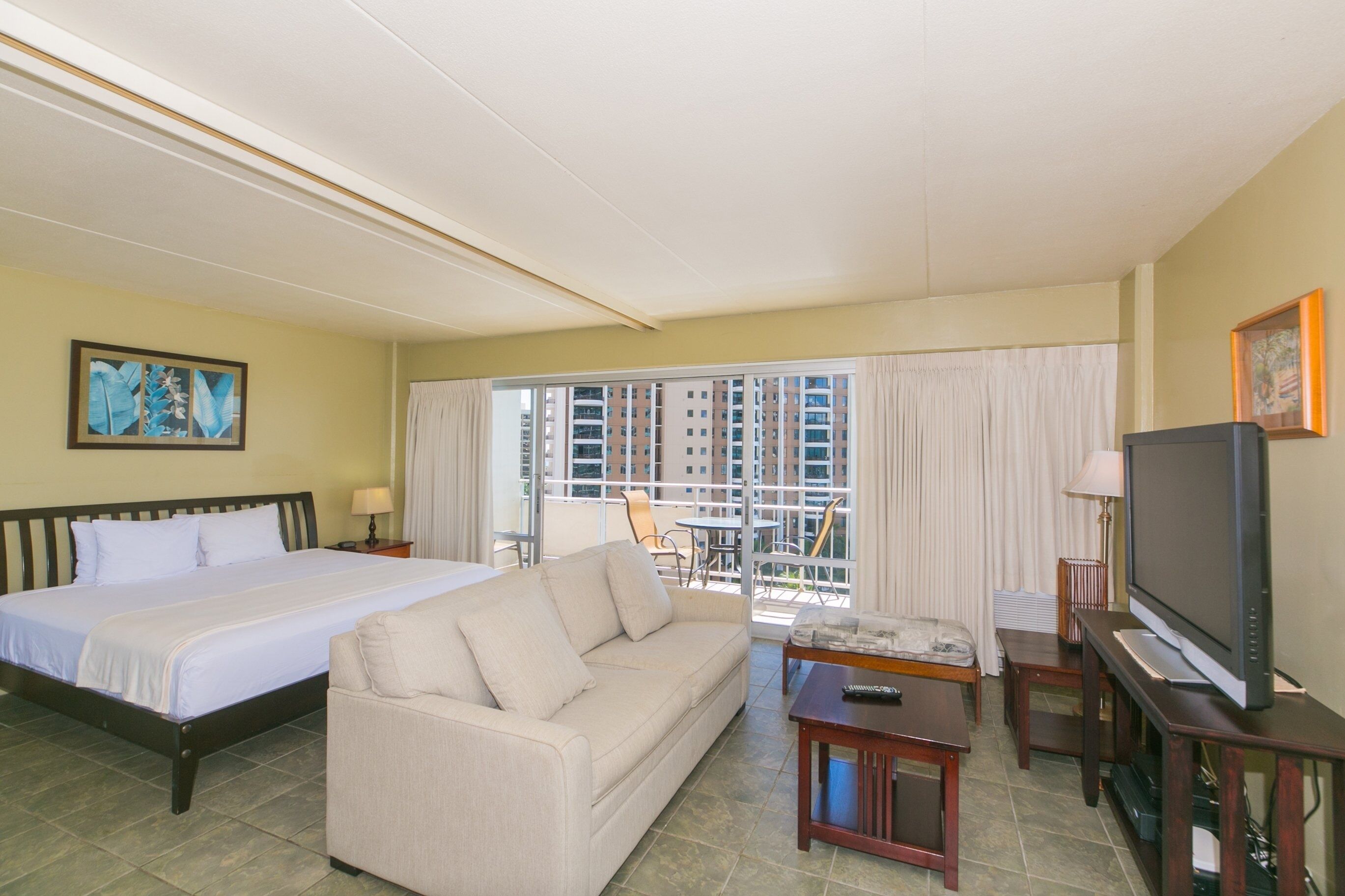 A hotel room with a large bed, a sofa, a television, and a balcony with a view of high-rise buildings.