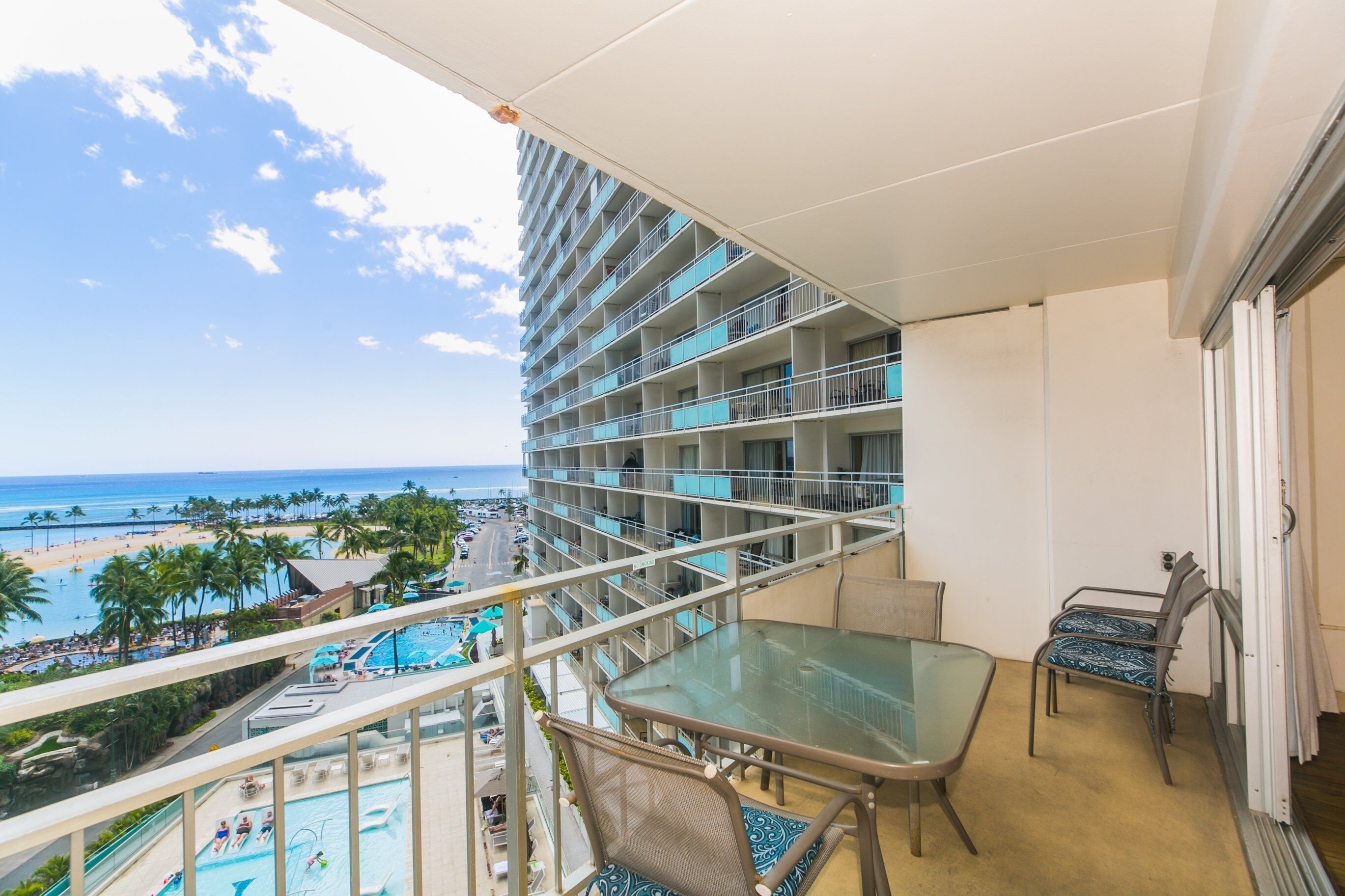 A balcony with a glass table and chairs, overlooking a pool and beach area.