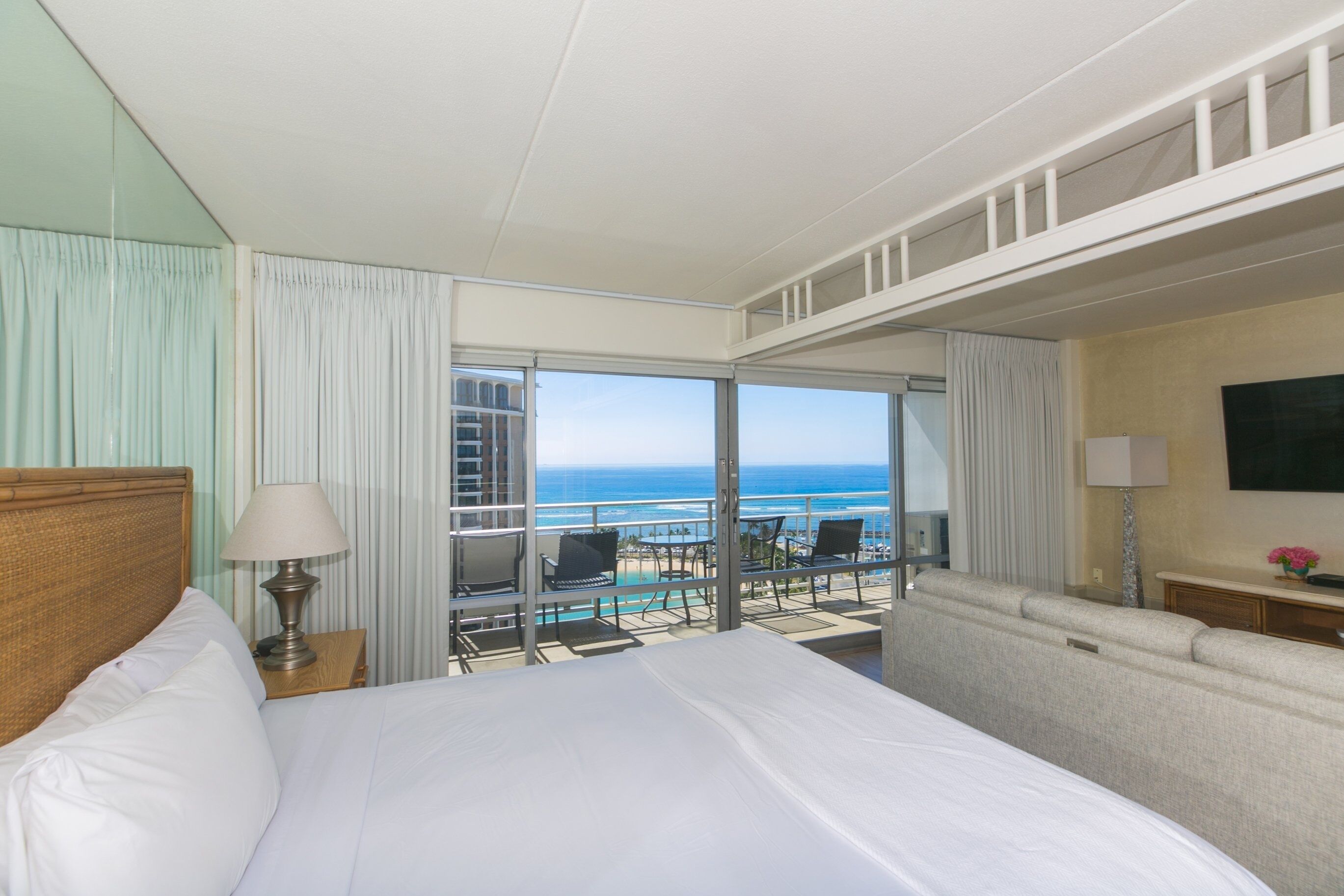 A hotel room with a bed, a nightstand, a lamp, a sofa, a TV, and a balcony with a view of the ocean.