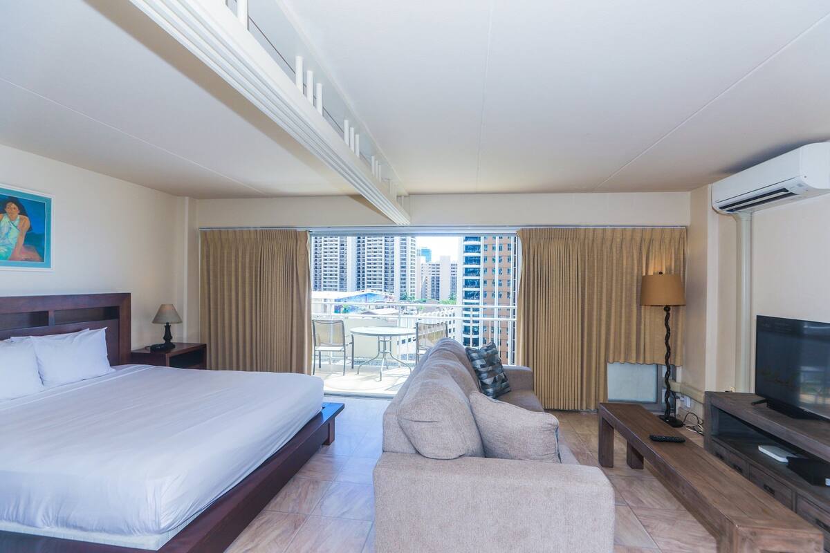 A hotel room with a large bed, a sofa, a TV, and a balcony with a view of high-rise buildings.