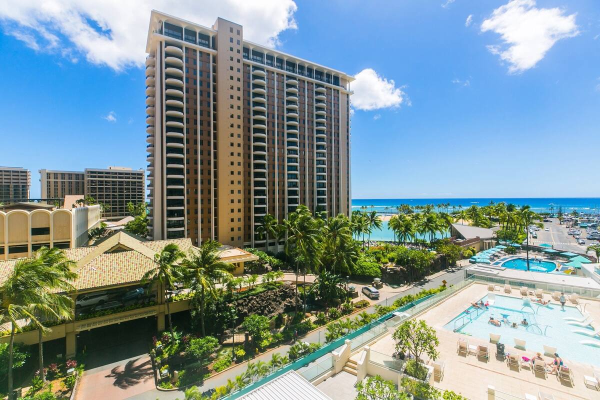 A high-rise hotel with a pool area, palm trees, and a view of the ocean.
