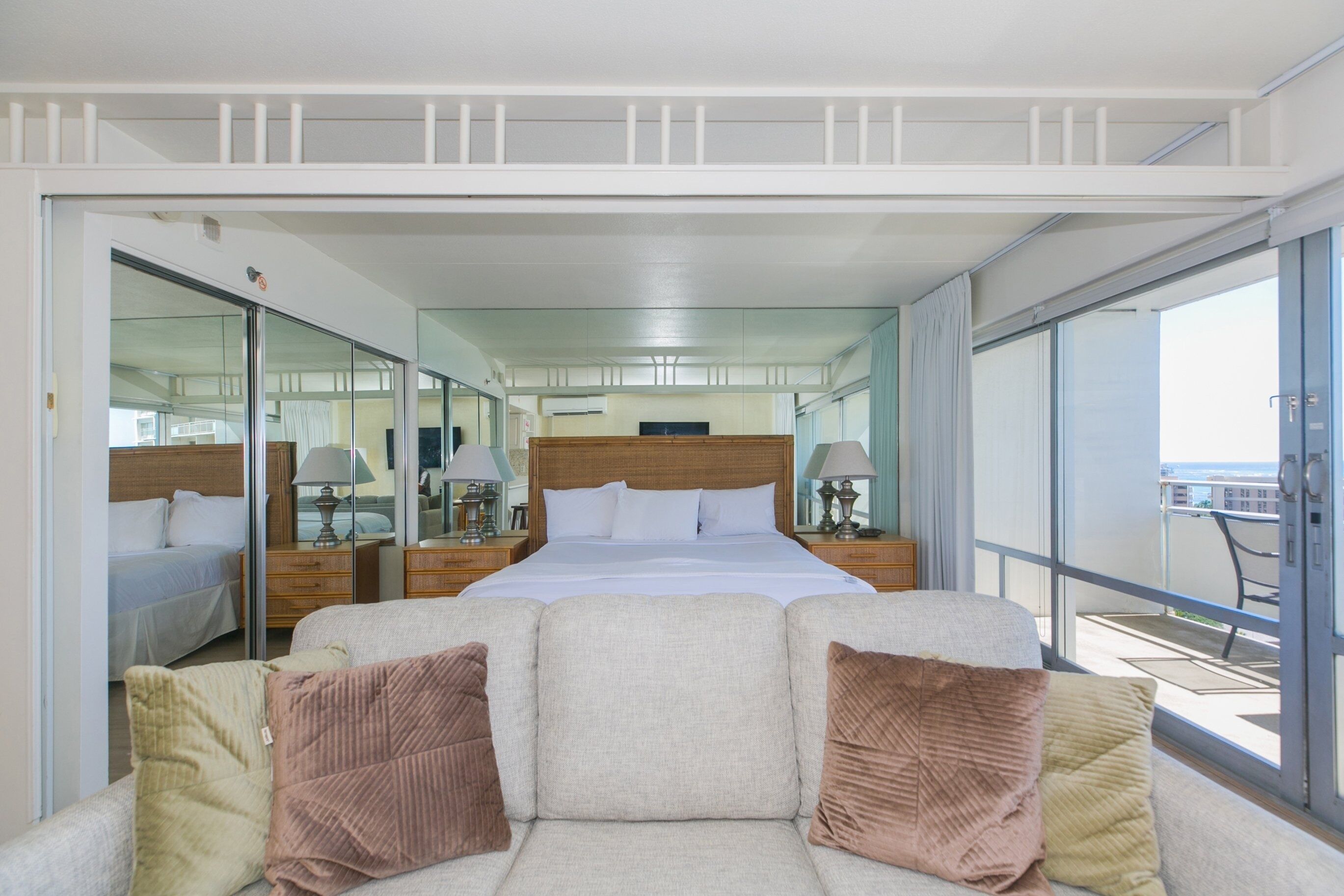 Une chambre d’hôtel moderne dotée d’un grand lit, d’un canapé avec des coussins, d’un balcon offrant une vue agréable et d’un grand miroir qui reflète la pièce.