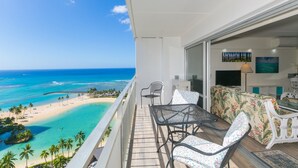 A balcony with a view of a beach and ocean, a dining table with chairs, and a sofa with a tropical print. - Ilikai Tower One Bedroom Lagoon View Waikiki Condos With Lanai & Free Wifi (Honolulu)