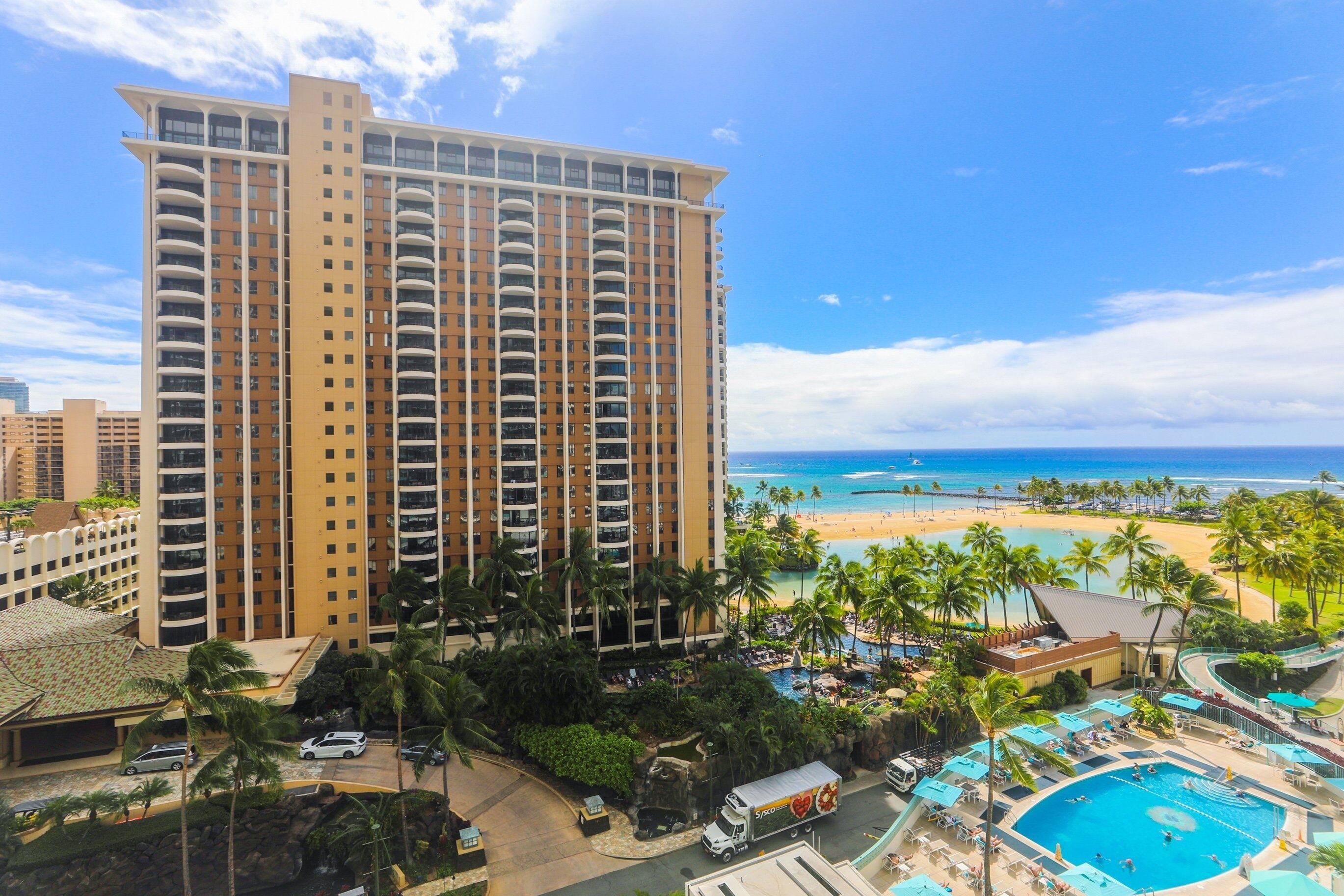 Un hotel de altura con zona de piscina, vista a la playa y palmeras.