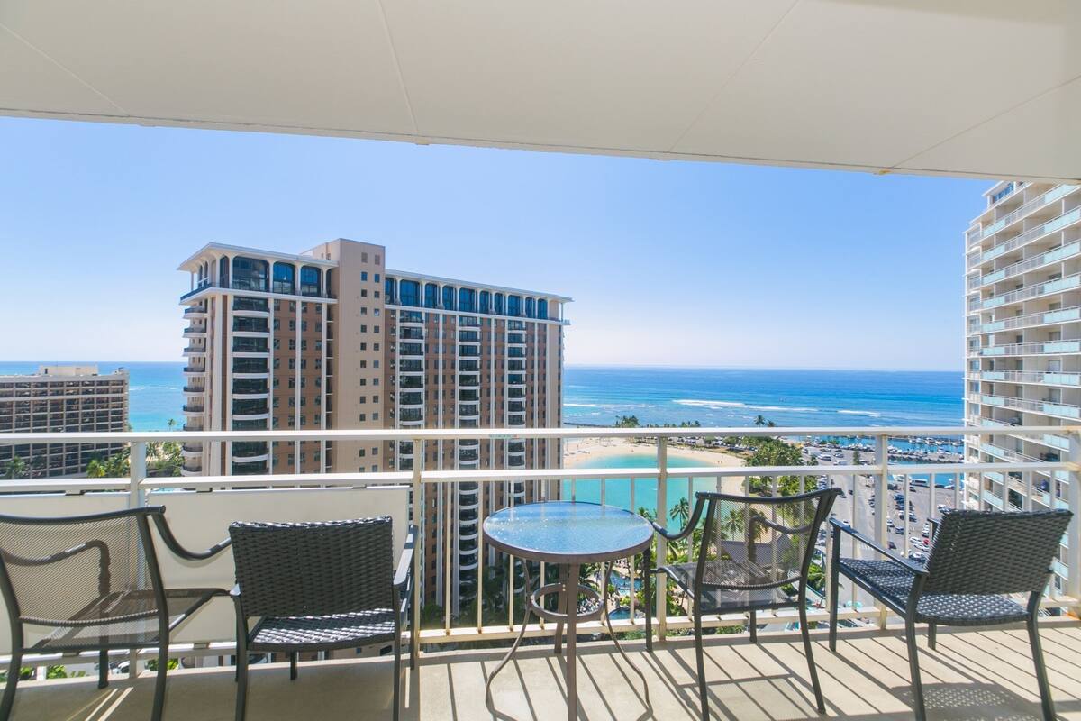 A balcony with a glass table and chairs, overlooking a beach and ocean.