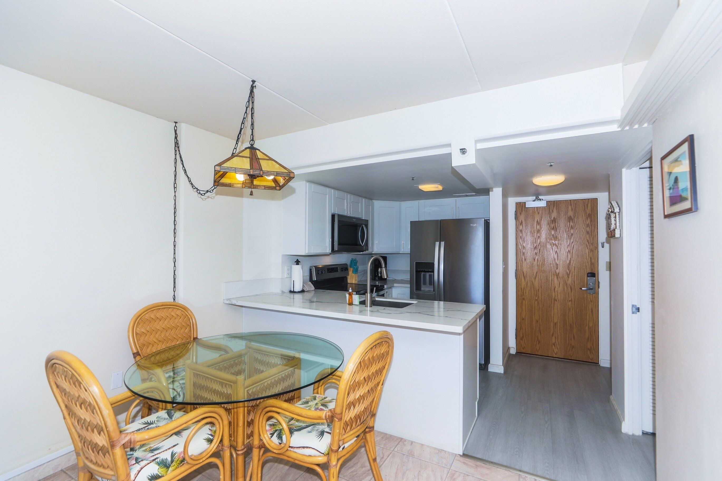 A compact kitchen with modern appliances, a dining area with a glass table and wicker chairs, and a hallway leading to a wooden door.