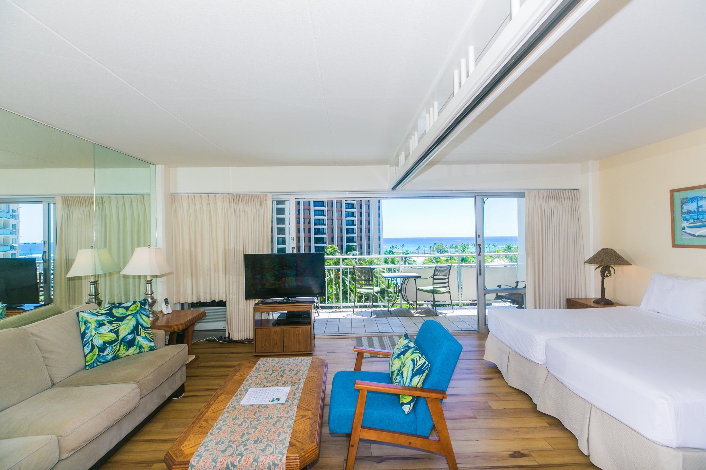 A hotel room with a sofa, a chair, a bed, a TV, and a balcony with a view of the ocean.