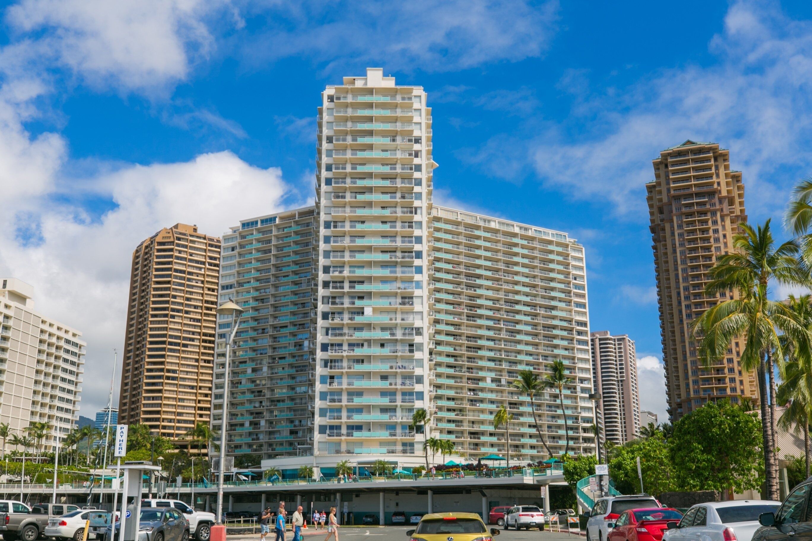 A high-rise hotel with multiple floors, a parking area, and palm trees in the foreground.