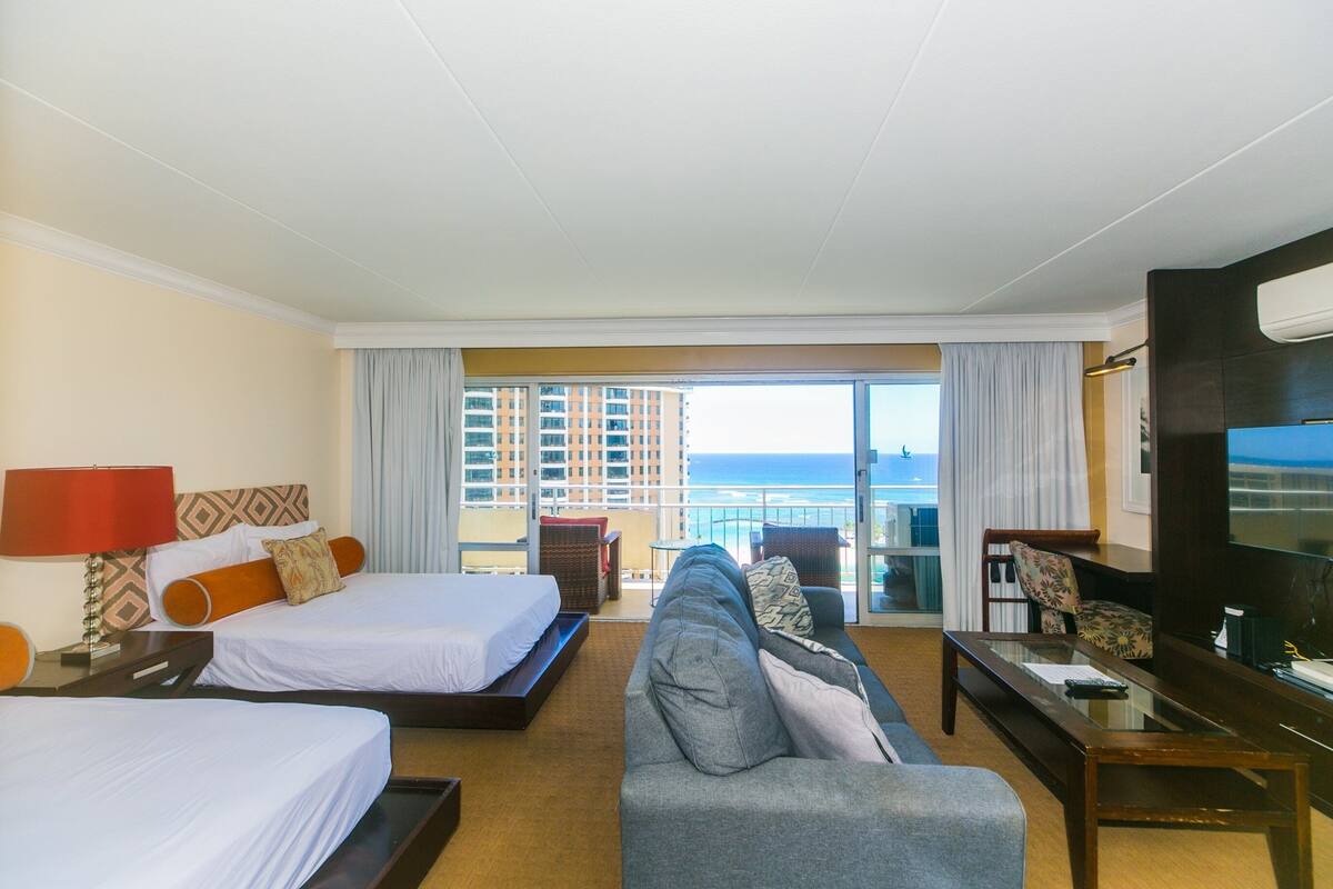 A hotel room with two beds, a sofa, a TV, and a balcony with a view of the ocean.