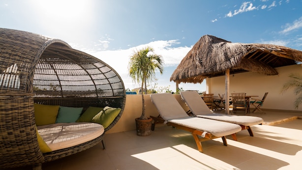 A sunlit outdoor seating area with wicker chairs and a thatched umbrella, a palm tree, and a tiled floor. - Condominio La Dolce Vita (Playa del Carmen)