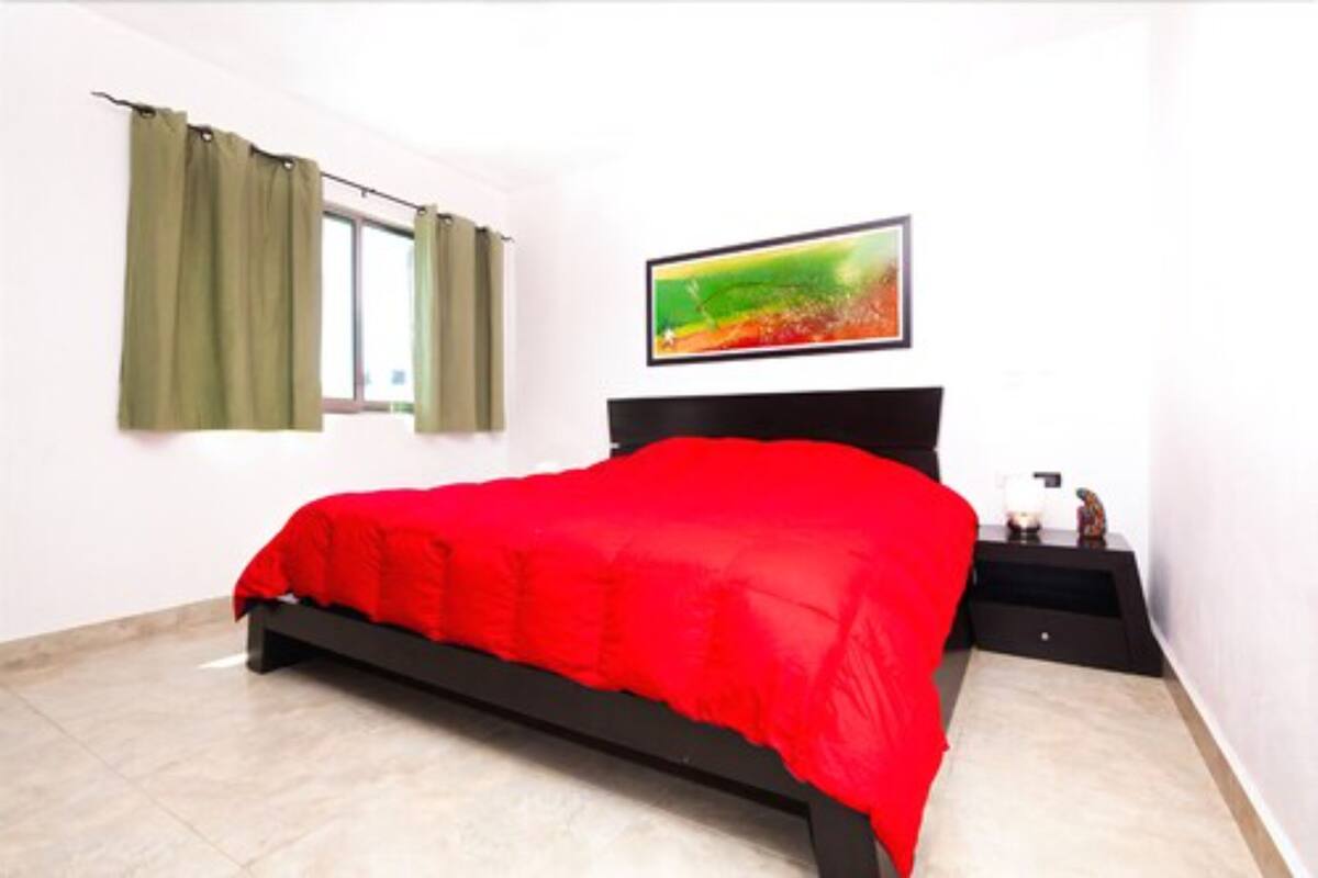 A bedroom with a bed, a window with curtains, a bedside table, and a wall-mounted TV.
