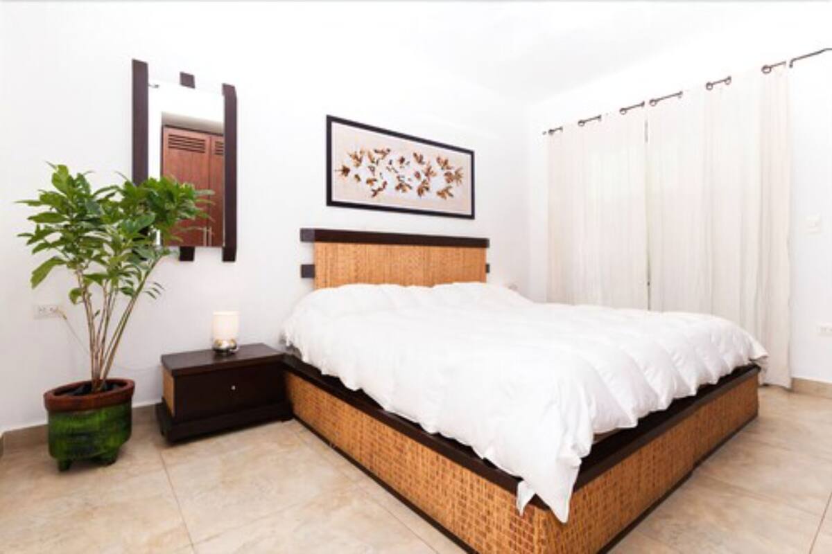 A bedroom with a bed, a bedside table, a potted plant, a framed picture, a mirror, and a white curtain.