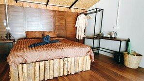Un dormitorio con cabecera de madera, una cama con colcha a rayas, un perchero, una mesita con una tetera y una cesta de mimbre.