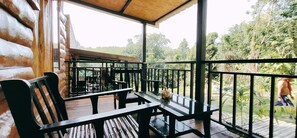 Um deck de madeira com mesas e cadeiras, ladeado por um guarda-corpo de metal, com vista para uma paisagem verde e exuberante.