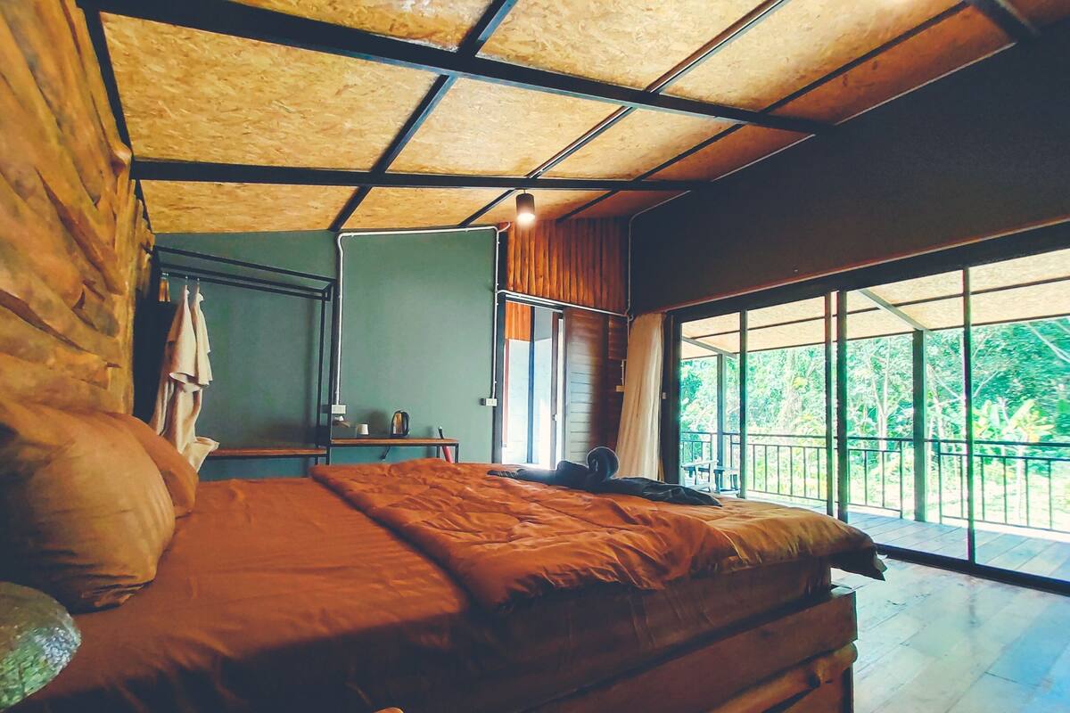 Una habitación con cama, una mesita, un espejo y ventana con vista a follaje.
