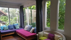 Una habitación con suelo de madera, una cama con colchón morado y almohadas azules, una silla de mimbre con cojines y ventanas amplias con cortinas.