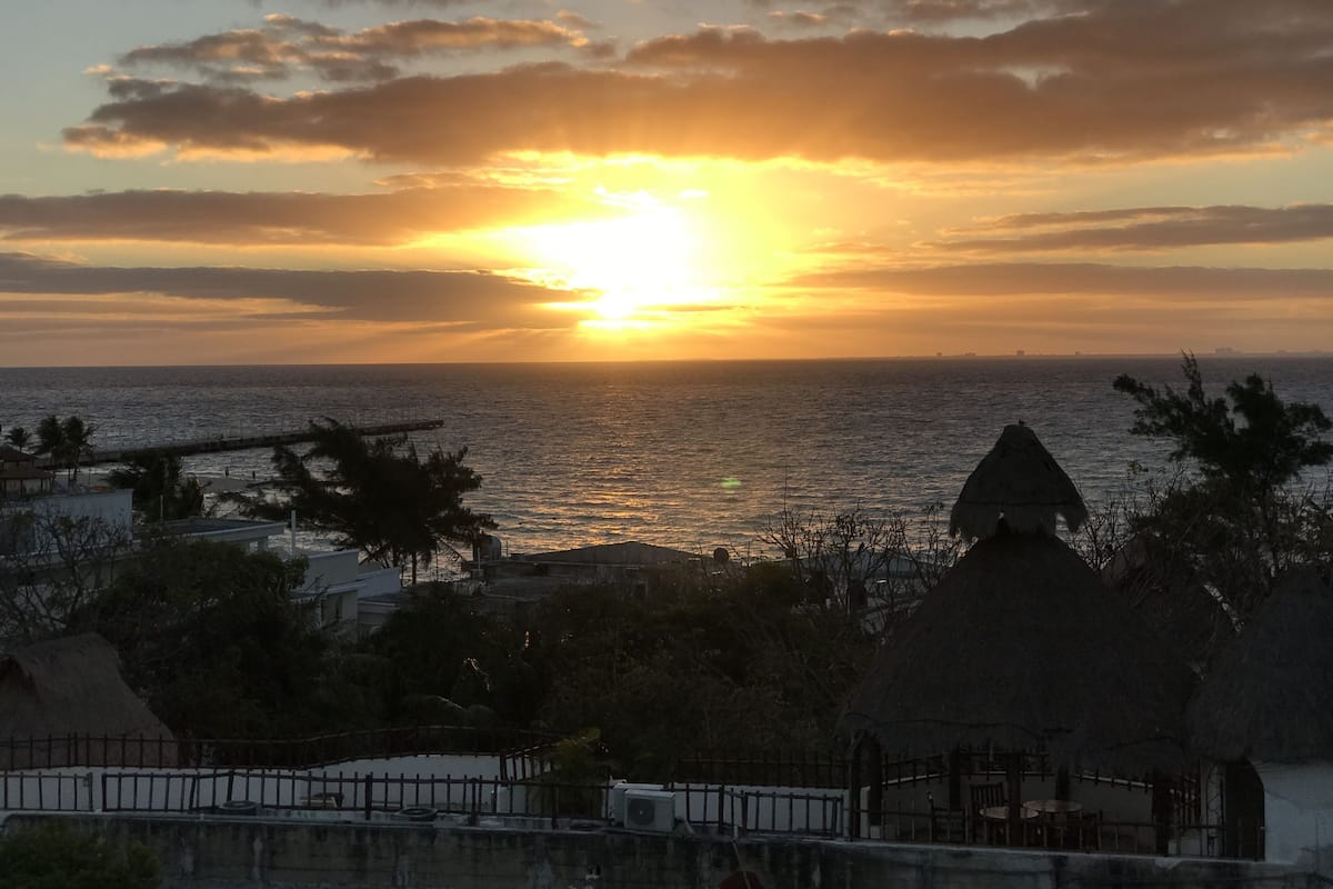 Un atardecer sobre el océano con un muelle y algunas cabañas con techo de paja en primer plano.